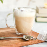 A foamy, lavender-sprinkled latte made with Coco-Lavender Chai in a glass mug, with an Heirloom Heart-Shaped Pewter Spoon full of Coco-Lavender Chai beside it. Both are on top of a wooden cutting board, with a stack of books in the distance.