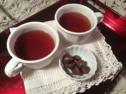 Two white teacups full of tea and a white dish of chocolates sit on a lacey white cloth on a red enamel tray.