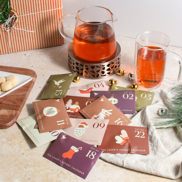 A colorful collection of Advent Calendar envelopes, surrounded by a teapot on a tea warmer, a mug of tea, a tea towel, some greenery, ornaments, shortbread cookies on a tray, and a festive gift.