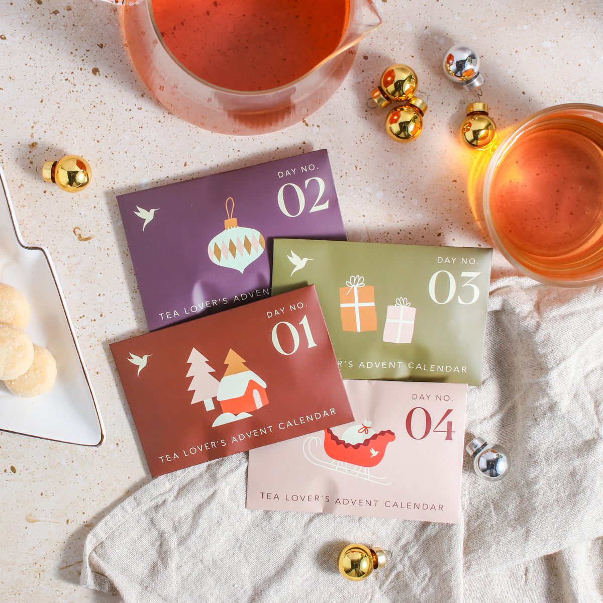 A colorful collection of 4 daily Advent envelopes, surrounded by silver and gold ornaments, shortbread cookies, a tea towel, a teapot, and a cup of tea shown from overhead.
