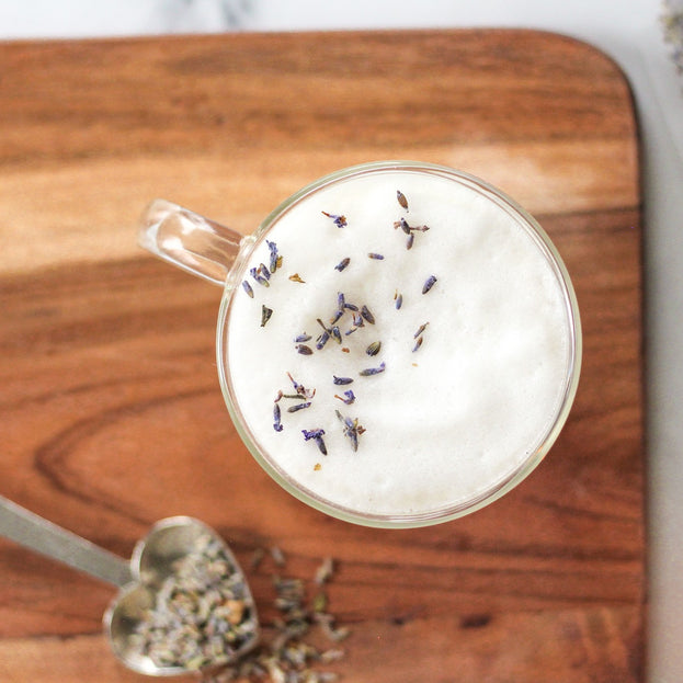 An overhead shot of a foamy, lavender-sprinkled latte made with Coco-Lavender Chai in a glass mug, with an Heirloom Heart-Shaped Pewter Spoon full of Coco-Lavender Chai beside it. Both are on top of a wooden cutting board.