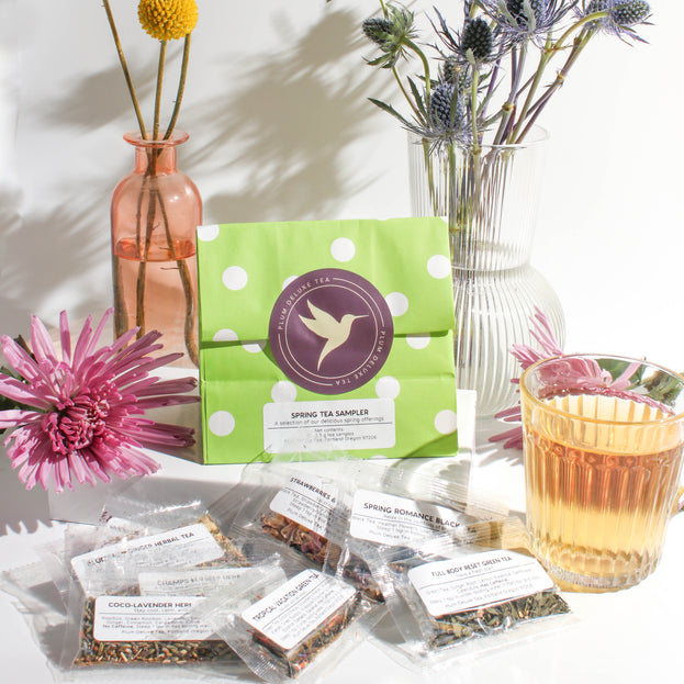 A green bag of tea samples, sealed with a purple hummingbird sticker, sits on a white block, surrounded by flowers in colorful vases, a glass of tea, and an assortment of tea samples, all on a white background.