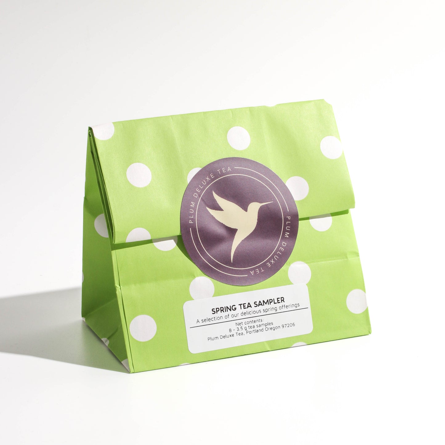 A green bag of spring tea samples, sealed with a purple hummingbird sticker, on a white background